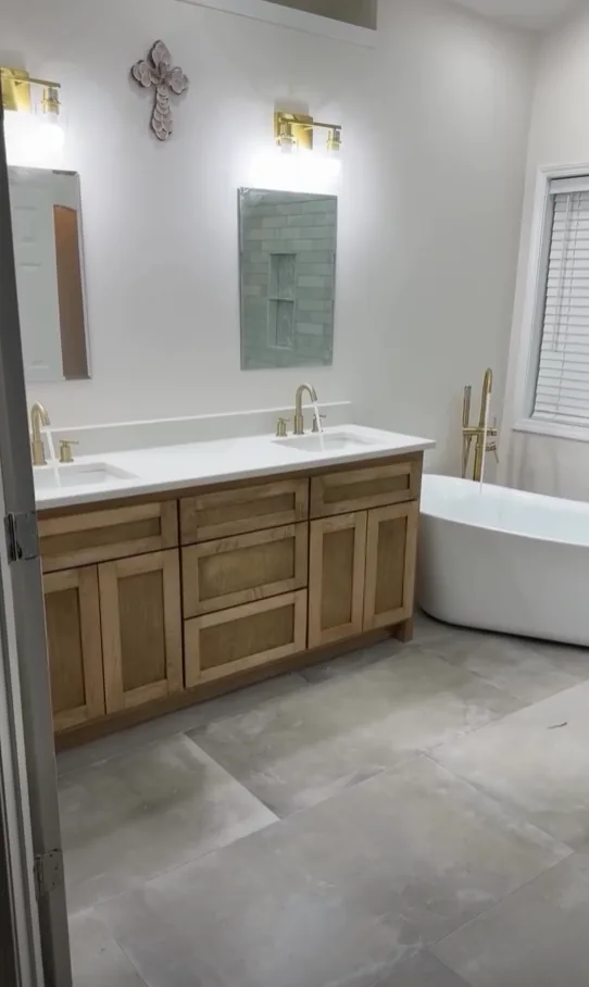 Double wood vanity with a white quartz top and two undermount sinks, brushed gold faucets and sconces, beside a freestanding soaking tub with a brushed gold floor-mounted filler on large-format gray floor tile