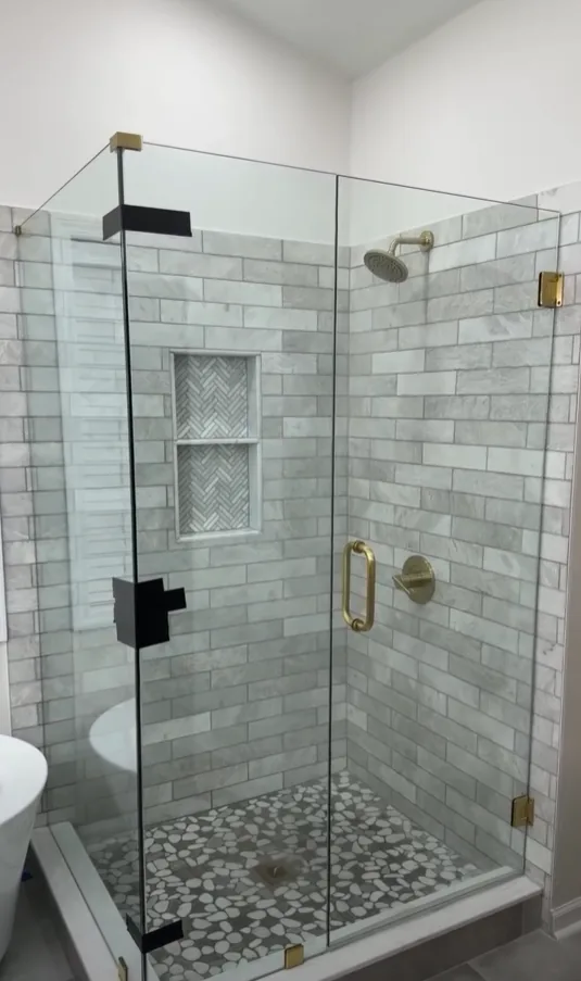 Frameless glass corner shower with brushed gold hinges and handle, marble-look subway tile walls, a two-shelf herringbone mosaic niche and a gray pebble tile floor