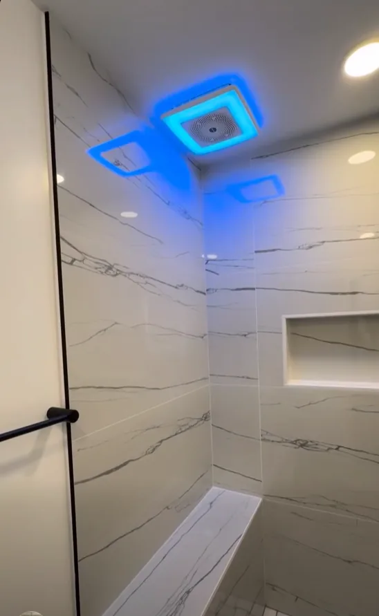 Inside a marble-look slab tile shower showing the tiled corner bench, a recessed niche and a blue-lit LED exhaust fan set into the ceiling