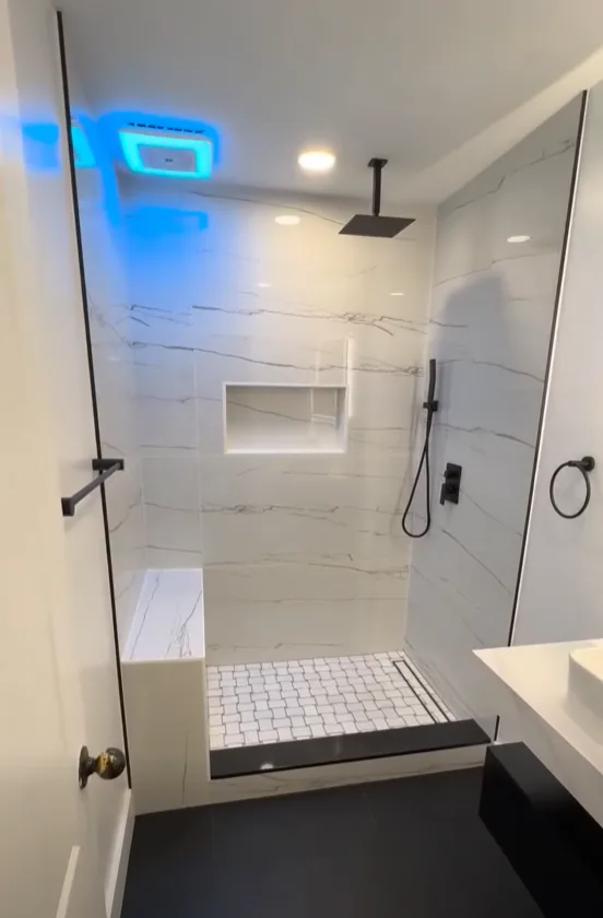 Walk-in shower tiled in white marble-look slab panels with a built-in bench, recessed niche, matte black ceiling-mounted rain head and handheld, octagon mosaic floor and a black linear drain