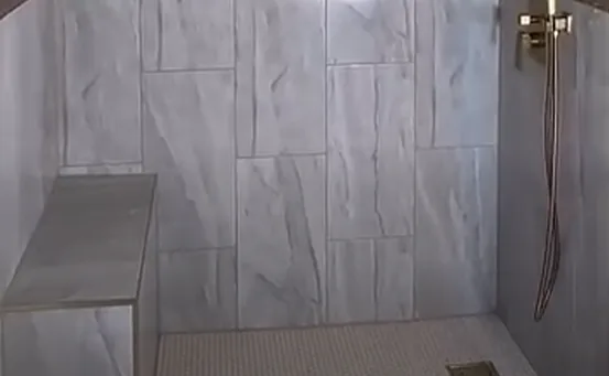 Tiled corner bench and a brushed gold handheld sprayer on a slide bar in a gray marble-look tile shower with a small mosaic tile floor